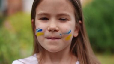 Headshot of patriotic charming Ukrainian girl with face painting looking at camera and looking away. Brunette child with brown eyes posing in slow motion outdoors on summer day