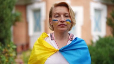 Confident Ukrainian woman gesturing peace standing on backyard in Ukrainian flag with blue and yellow face painting. Brave lady posing in slow motion outdoors. Hope and patriotism concept