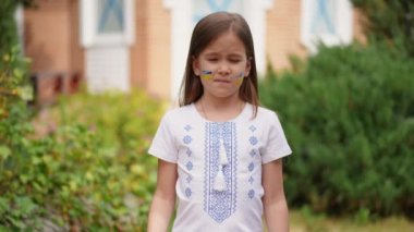 Sad girl in traditional clothing looking at camera with serious facial expression showing Help Ukrainian children placard. Portrait of depressed frustrated child posing in slow motion asking support