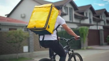 Tracking shot of young Caucasian man with yellow delivery bag riding bicycle on suburban street. Live camera follows delivery boy delivering food and drink on summer day outdoors. Service concept