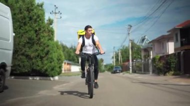 Wide shot front view confident delivery boy driving bicycle fast on sunny street in town. Caucasian handsome man delivering order outdoors in sunshine riding bike. Speed and service