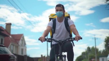Front view portrait of young man in coronavirus face mask riding bicycle delivering food and drink in urban city. Confident Caucasian delivery boy driving bike with backpack at background of sky