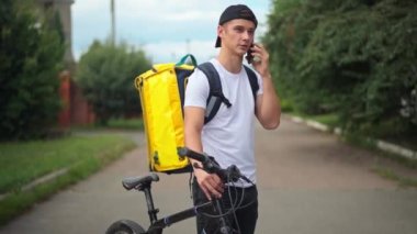 Caucasian courier talking on phone with client standing on suburban road with bike and yellow delivery bag. Portrait of handsome young delivery boy searching customer in town