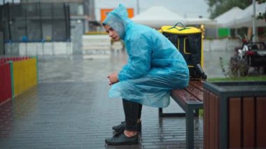 Wide shot side view of sad young courier sitting on rainy day in city waiting. Upset Caucasian delivery boy in rain coat with backpack outdoors in urban town. Lifestyle and profession concept