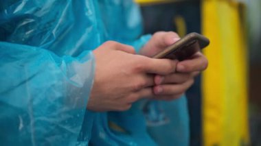 Close-up male hands typing on smartphone touchscreen outdoors. Unrecognizable Caucasian courier in rain coat messaging online with yellow delivery bag at background