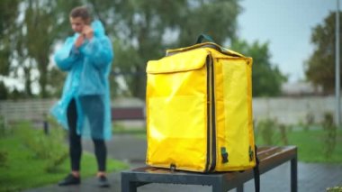 Unrecognizable blurred delivery man hanging up phone leaving with yellow backpack in urban city park. Young Caucasian courier in rain coat walking away with food delivery bag