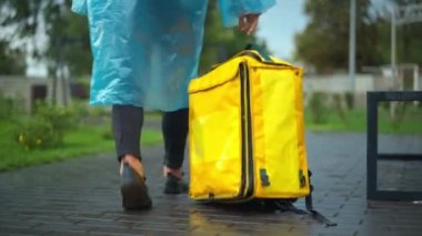 Unrecognizable young man in rain coat leaving yellow delivery backpack walking away in city park. Stressed Caucasian male courier quitting part-time job. Work and lifestyle