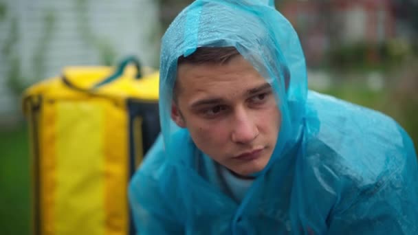 Close Portrait Depressed Young Man Rain Coat Delivery Backpack Sitting — Vídeo de stock