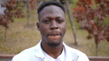Headshot portrait of cheerful African American young man with toothy smile looking at camera posing in slow motion outdoors. Close-up happy carefree positive guy smiling in autumn park outdoors
