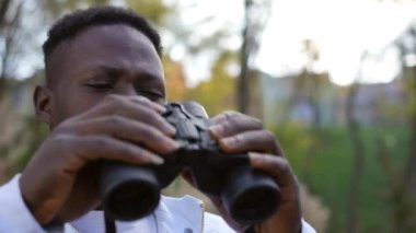 African American young man using binoculars looking away standing in forest on autumn day. Close-up portrait of curios handsome traveler enjoying eco-tourism outdoors in forest