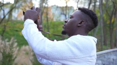 Side view concentrated talented African American photographer taking photos of autumn nature in park. Inspired young man photographing outdoors admiring beauty of nature