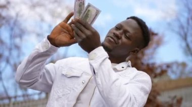 Satisfied smiling man scattering money standing in autumn city park. Portrait of wealthy rich African American young guy enjoying wealth outdoors