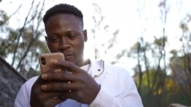 Portrait of smiling man in sunrays reading message on smartphone standing outdoors. Happy African American guy with toothy smile in sunbeam in autumn park with phone