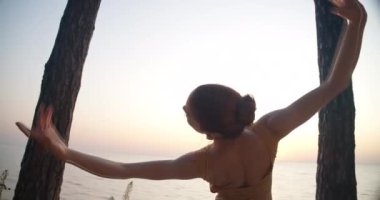 Live camera spinning as graceful ballerina dancing between trees at background of sunrise sunshine. Slim talented woman moving hands performing on sea shore outdoors. Cinema 4k ProRes HQ