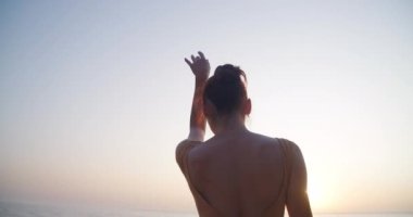 Live camera follows movement of slim graceful ballerina dancing at sunset on sea shore in slow motion. Back view young Caucasian talented confident woman performing in sunrays. Cinema 4k ProRes HQ