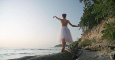 Back view elegant slim woman in tutu standing on tree trunk on river bank moving hands in slow motion. Wide shot graceful Caucasian ballet dancer performing outdoors at sunset. Cinema 4k ProRes HQ