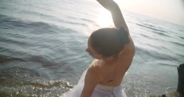 Back view slim ballerina dancing in sunrays at background of waves with live camera following movement in slow motion. Graceful confident Caucasian woman rehearsing in sunbeam. Cinema 4k ProRes HQ