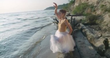 Portrait of gorgeous flexible ballet dancer moving hands bending sitting on tree trunk on sea shore. Beautiful Caucasian ballerina performing rehearsing outdoors at sunrise. Cinema 4k ProRes HQ