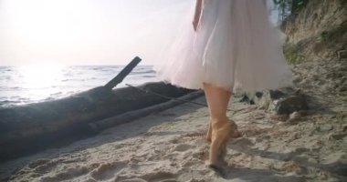 Tracking shot of slim graceful woman in pointies walking on sandy beach at dawn. Caucasian talented elegant ballerina in tutu strolling in slow motion on river bank at sunrise. Cinema 4k ProRes HQ