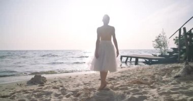Back view slim ballerina walking in sunbeam on sandy beach at sunset. Young talented Caucasian woman in tutu making steps in slow motion strolling to waves rolling on sand. Cinema 4k ProRes HQ