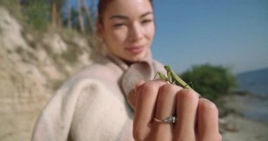 Close-up green mantis on female Caucasian hand with blurred young woman smiling at background. Insect in sunshine outdoors with confident lady admiring mantid. Cinema 4k ProRes HQ