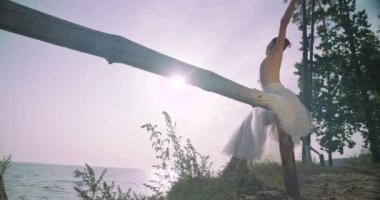 Young slim woman in tutu sitting on wooden fence on the right in sunshine moving hands in slow motion. Side view portrait of Caucasian ballet dancer rehearsing at sunrise. Cinema 4k ProRes HQ
