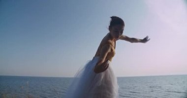 Slim ballerina dancing at background of blue sea and clear summer sky outdoors. Back view portrait of Caucasian graceful talented ballet dancer performing in slow motion at dawn. Cinema 4k ProRes HQ