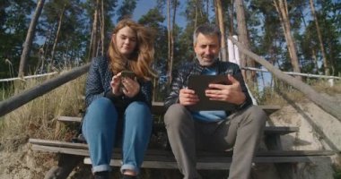 Front view young smiling Caucasian woman scrolling smartphone as positive mature man talking showing digital tablet. Portrait of happy carefree relaxed father and daughter outdoors. Cinema4k ProResHQ