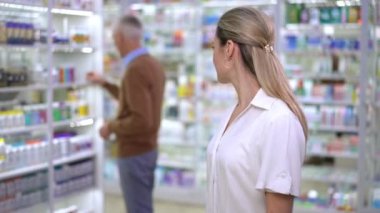 Positive smiling woman in medical gown on the right looking back at senior man choosing pills turning looking at camera. Portrait of confident professional pharmacist posing in drugstore with client