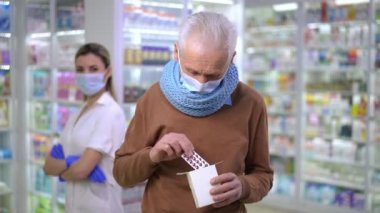 Portrait of happy senior man advertising pills standing in Covid-19 face mask in pharmacy with blurred woman with crossed hands at background. Satisfied Caucasian client posing with efficient drug
