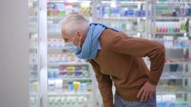 Side view senior Caucasian man leaning at shelves in pharmacy choosing efficient medication on coronavirus pandemic. Ill grey-haired client examining drugs indoors thinking. Medicine concept