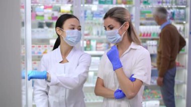 Young professional Asian and Caucasian women discussing efficiency of drugs with blurred client choosing pills at background in pharmacy. Pharmacists in coronavirus face masks talking in drugstore