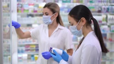 Side view Asian female pharmacist collecting expired drugs on shelves leaving with Caucasian colleague. Young beautiful women working in drugstore walking away with medication bottles