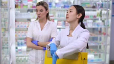 Overburdened Asian young pharmacy assistant with box standing at shelves with angry Caucasian manager arguing at background. Sad new employee making mistake in pharmacy. Professionalism concept