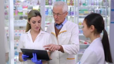 Expert senior pharmacist talking with young Caucasian and Asian employees in pharmacy explaining drugs variety. Portrait of professional man discussing medications with new colleagues in drugstore