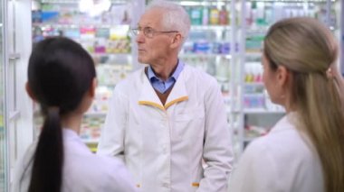 Senior man in eyeglasses talking with new employees standing at shelves in pharmacy. Portrait of confident intelligent pharmacist explaining drugstore management to young Asian and Caucasian women