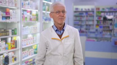 Portrait of confident senior pharmacist crossing hands looking at camera smiling. Medium shot of grey-haired Caucasian intelligent man in eyeglasses posing in drugstore. Health care and intelligence