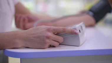 Close-up electronic sphygmomanometer with female hand pressing button and blurred arm with blood pressure gauge at background. Unrecognizable doctor and patient in hospital indoors