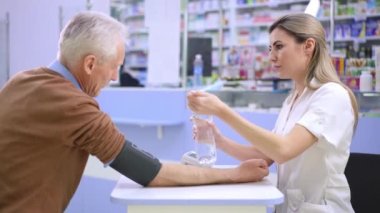 Side view smiling caring female pharmacist passing water for unwell senior man taking off blood pressure gauge. Portrait of intelligent professional Caucasian woman supporting client in drugstore