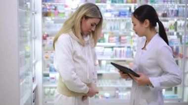 Pregnant woman putting on maternity belt talking with pharmacist in drugstore. Beautiful Caucasian and Asian women choosing belly belt in pharmacy standing with digital tablet