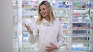 Portrait of happy pregnant beautiful woman advertising pills standing in pharmacy indoors. Front view of smiling Caucasian expectant looking at camera stretching bottle with medication