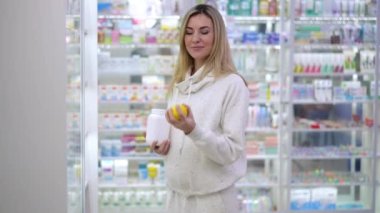 Charming smiling pregnant woman advertising multivitamin complex in pharmacy juggling lemon fruit. Portrait of gorgeous Caucasian expectant looking at camera standing in drugstore with pills bottle