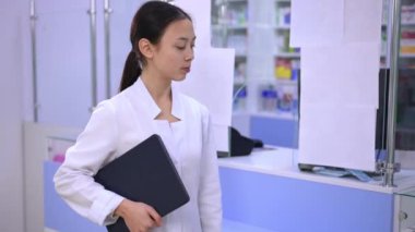 Female Asian pharmacist cashier posing with digital tablet at cash desk in drugstore. Portrait of positive confident beautiful young woman looking at camera smiling standing in pharmacy indoors