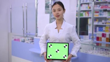 Young Asian woman posing with green screen digital tablet at cash desk in pharmacy. Portrait of positive intelligent pharmacist smiling looking at camera standing with chromakey web page template