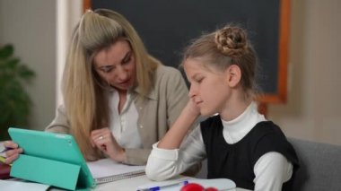 Side view tired teenage girl sitting with teacher at desk talking. Exhausted Caucasian student learning as blurred woman explaining in classroom in school. One-on-one education and knowledge