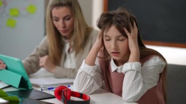 Exhausted overburdened teen schoolgirl looking at camera sighing as blurred teacher talking at background. Portrait of tired sad Caucasian girl posing in classroom with woman explaining lesson