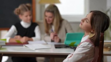 Sad bored schoolgirl crossing hands sighing looking at camera with dissatisfied facial expression as nerd classmate and teacher talking at background. Portrait of Caucasian girl posing on the right