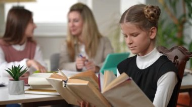 Genius teenage girl reading books sitting in classroom with blurred classmate and teacher and looking at camera smiling. Portrait of smart intelligent Caucasian student posing in school indoors