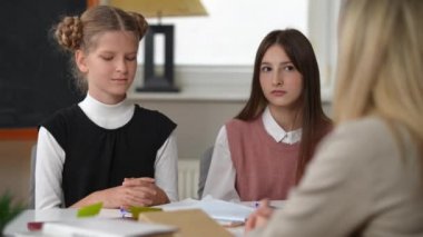 Stressed Caucasian teen schoolgirl leaving classroom as teacher scolding student. Portrait of dissatisfied cute girl walking away as classmate and tutor sitting at desk in school. Education failures