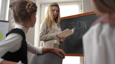 Smart positive teacher reading book explaining lesson talking with teen students in classroom. Portrait of confident intelligent Caucasian woman teaching teenage schoolgirls indoors smiling